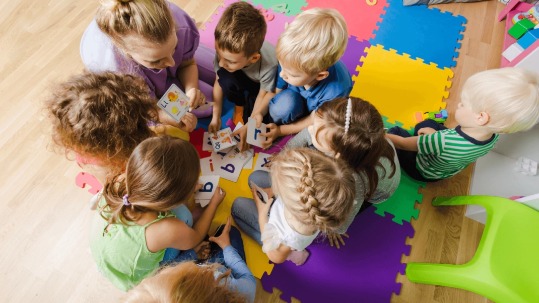 A group of young children sit on colorful foam mats, gathered around an adult, holding letter cards and engaging in a learning activity.